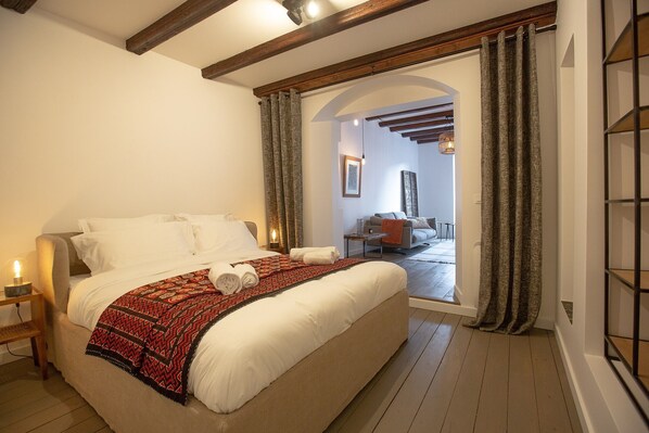 A bedroom with a large bed, a nightstand, and a view of a living area through an open door.