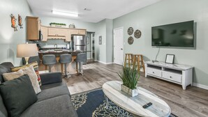 A modern living room with a sofa, coffee table, and TV. The room has wooden flooring, a ceiling fan, and a kitchen in the background. - 3 Bedroom Townhome 87B, one block to beach, close to casinos, & attractions (Gulfport)