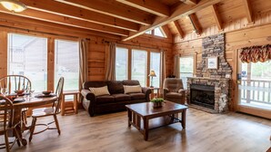 A log cabin interior with a dining area, a sofa, a fireplace, and large windows.