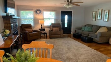 A living room with a fireplace, two armchairs, a sofa, a dining table with chairs, and a ceiling fan.