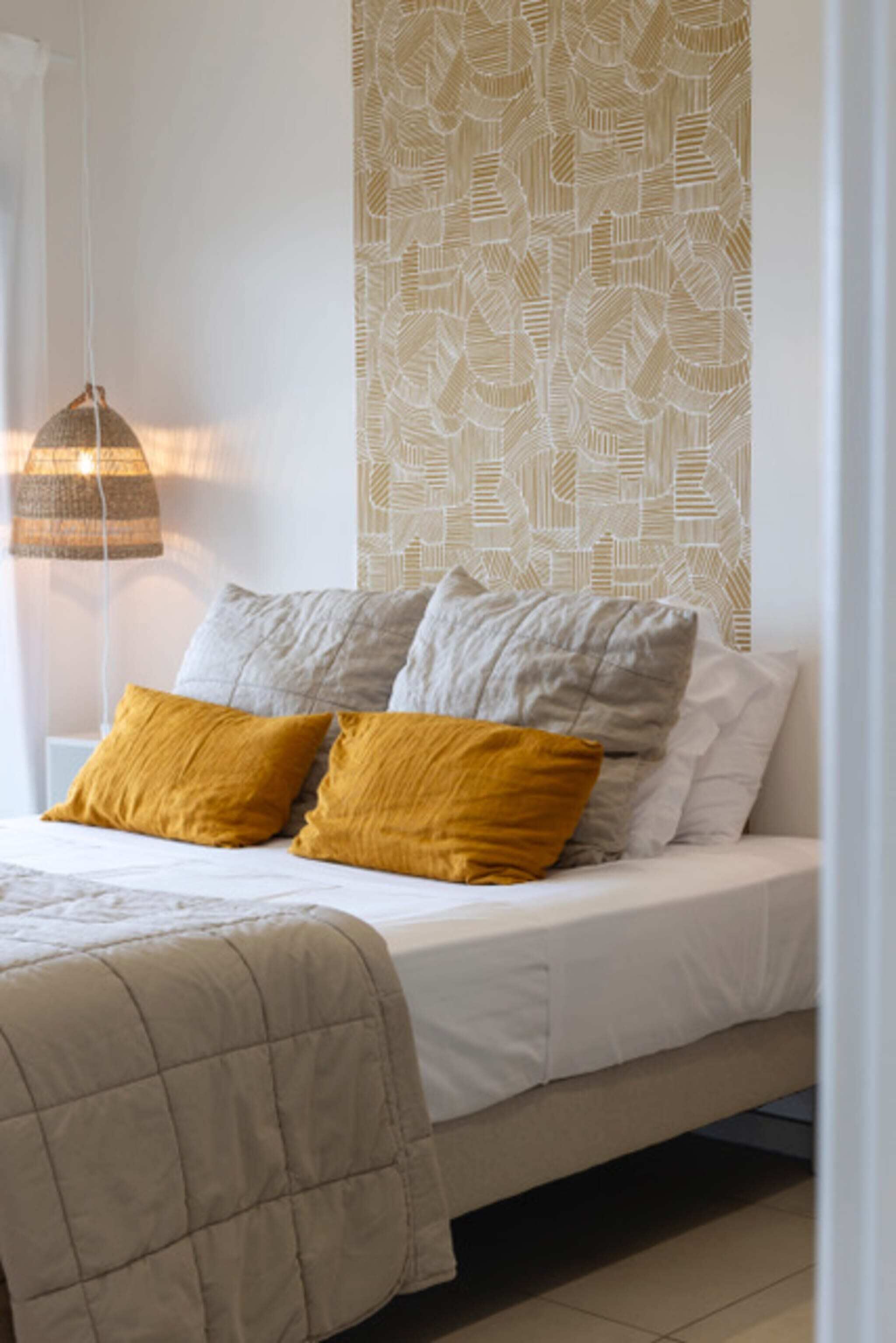 Un lit bien fait, recouvert d’une couette beige, avec deux oreillers de couleur moutarde et gris, et un mur à motifs en arrière-plan.