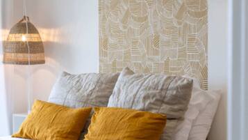 Un lit bien fait, recouvert d’une couette beige, avec deux oreillers de couleur moutarde et gris, et un mur à motifs en arrière-plan.
