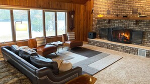 A cozy living room with a brick fireplace, leather sofas, and a large window overlooking a yard. - Your Northwoods Waterfront Getaway (Crivitz)