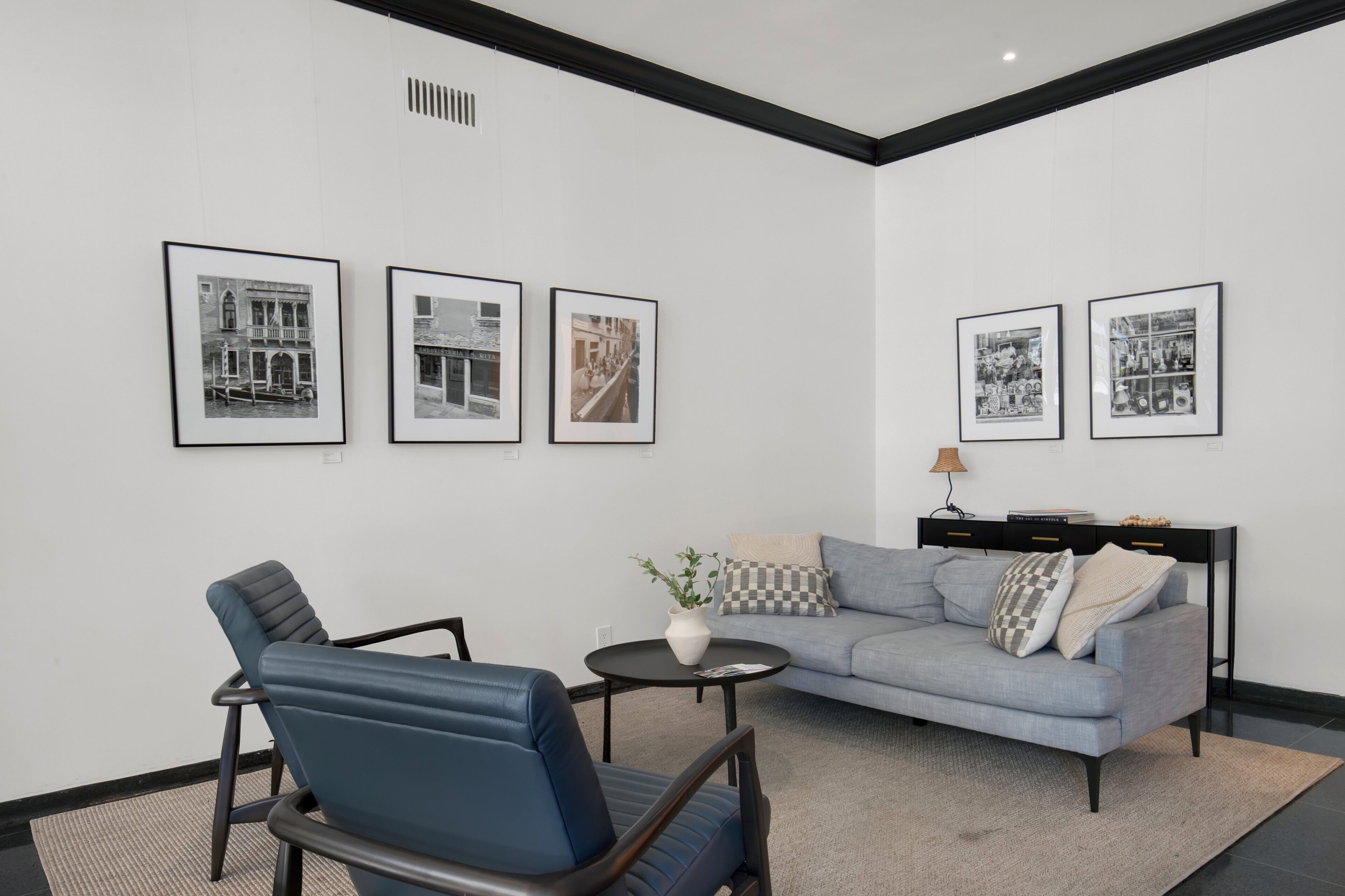 A modern room with a grey sofa, a blue chair, a small round table with a vase, and four framed pictures on the wall.