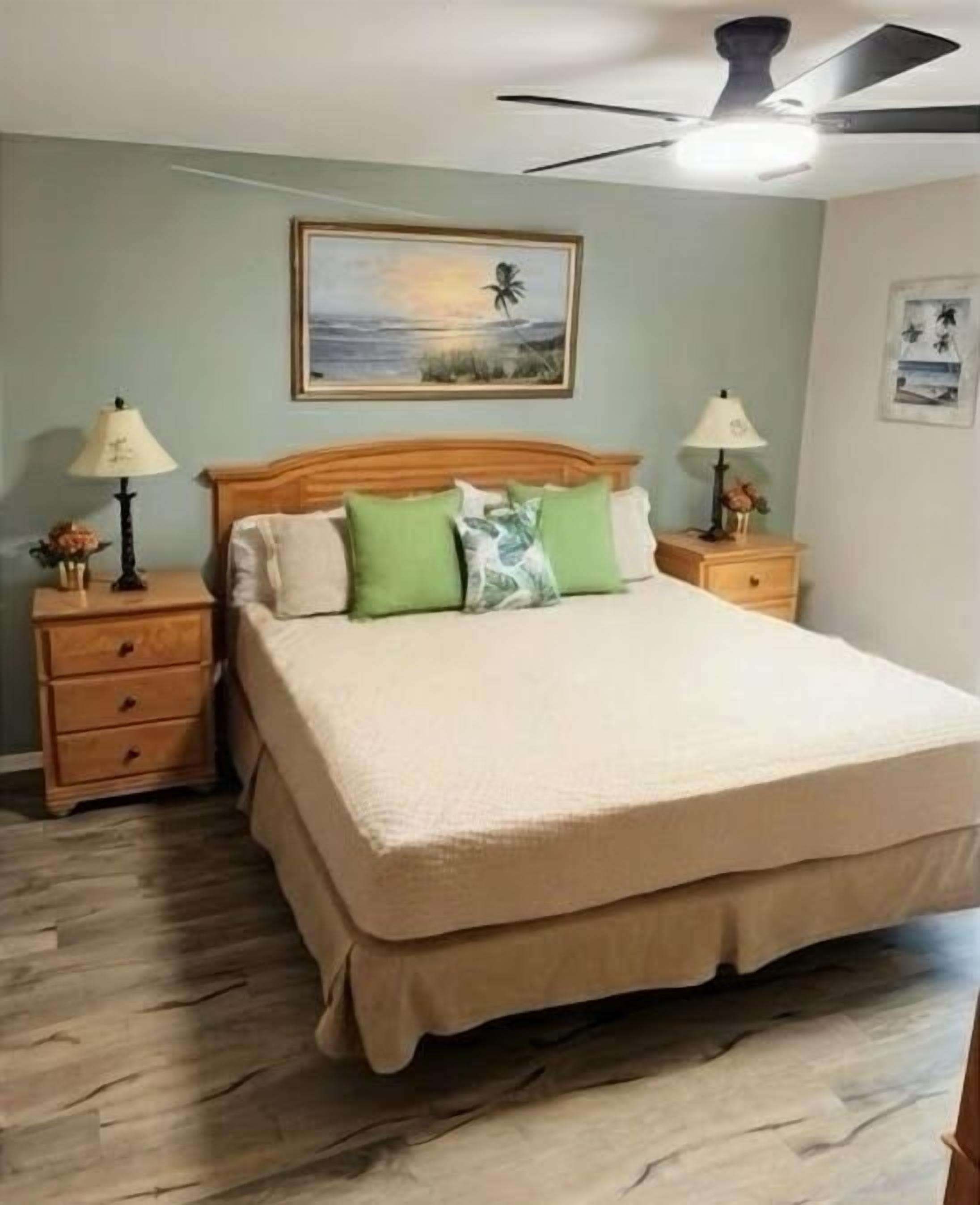A bedroom with a large bed, two nightstands, a ceiling fan, and a painting on the wall.