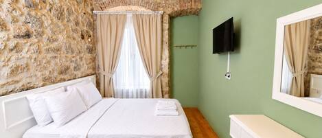 Un dormitorio con pared de piedra, cama con ropa de cama blanca, techo de madera, televisor montado en la pared y ventana con cortinas.