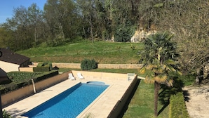 Una piscina rettangolare con due sdraio bianche, circondata da erba curata e palme, con un muro in pietra e un edificio sullo sfondo.