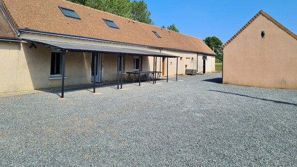 Un bâtiment avec un toit en tuiles, un espace extérieur couvert aménagé en coin salon et une allée gravillonnée.