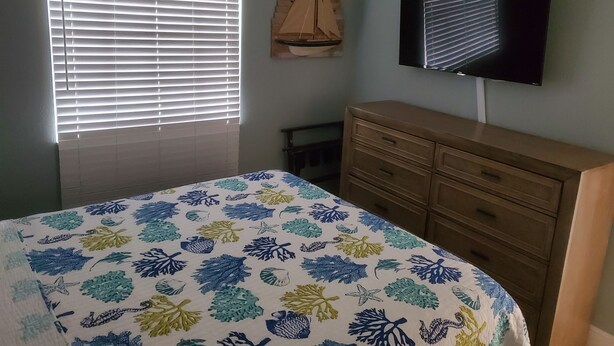 Un dormitorio con cama, cómoda, televisor en la pared y ventana con persianas.