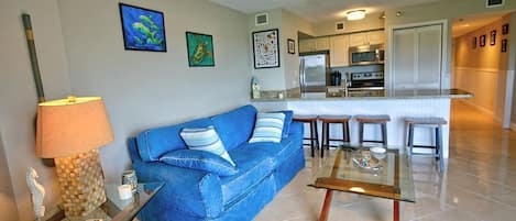 Una sala de estar con un sofá azul, una mesa de centro de vidrio y una lámpara. Se observa una cocina al fondo con un refrigerador, un microondas y una estufa.