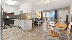 A modern kitchen with white cabinets, stainless steel appliances, and a wooden dining table with chairs. - Coastal Beach Condo (Carolina Beach)