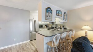 A modern kitchen with white cabinets, stainless steel appliances, and a granite countertop. There are three bar stools and a black couch in the dining area. - Coastal Beach Condo (Carolina Beach)