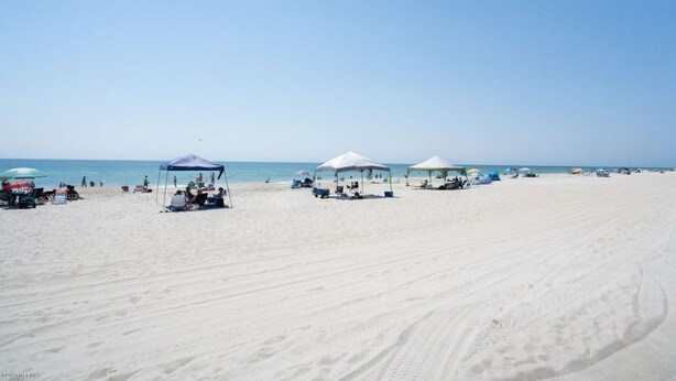 A beach with white sand, clear blue sky, and several beach umbrellas and tents. - Coastal Beach Condo (Carolina Beach)
