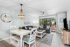 A modern living room with a dining area, a ceiling fan, and a large window with a view of greenery.
