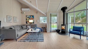 Ein gemütliches Wohnzimmer mit grauem Sofa, einem Couchtisch und einem blauen Sessel. Ein Kaminofen ist zu sehen, und große Fenster bieten einen Blick ins Freie.