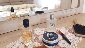 Un tavolino ordinato con uno specchio, una bottiglia di Acqua di Gio, una scatola Nivea Men, uno spazzola per capelli e un braccialetto dorato su una superficie in legno con un sottile ricamo in pizzo.
