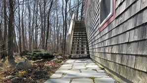 Ein steinbelagter Weg führt zu einer Holztreppe, begleitet von einem Holzzaun auf der einen Seite und einem Gebäude mit verkleideter Fassade auf der anderen.