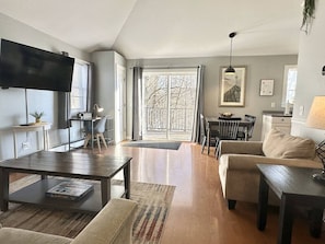 A living room with a TV, sofa, coffee table, and a dining area with chairs and a table. - 2BR Apt + Deck, Fire Pit, Backyard! [Maine Escape] (Bar Harbor)