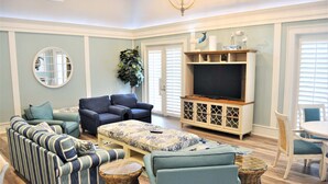 A living room with a striped sofa, a blue armchair, a patterned ottoman, wicker side tables, a television, and a dining area with a white table and chairs. - "Sunrise Sonata" Direct Oceanfront Penthouse Stunning Sunrises • Holiday Promo (Palm Coast)