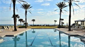 A swimming pool with clear blue water, surrounded by palm trees and lounge chairs, with a view of the ocean in the background. - "Sunrise Sonata" Direct Oceanfront Penthouse Stunning Sunrises • Holiday Promo (Palm Coast)