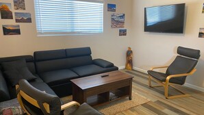 Una sala de estar con un sofá negro, sillas de madera, una mesa de centro y un televisor en la pared.