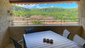 A balcony with a table and chairs, overlooking a village with terracotta roofs and lush green hills.