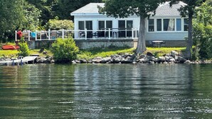 Una casa frente al lago, con un muelle, una zona de estar cubierta al aire libre y un gran árbol en primer plano.