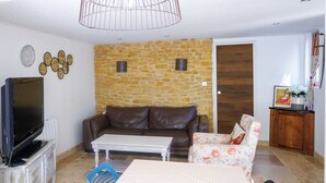 Una sala de estar con una pared de piedra, un sofá de cuero marrón, una mesa de centro blanca, un sillón estampado y un televisor sobre un mueble, además de una puerta de madera.