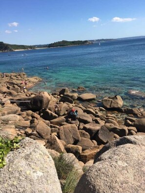 Une plage rocheuse avec une eau claire et des bateaux qui se dessinent à l’horizon.