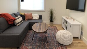 A modern living room with a grey sofa, a round wooden coffee table, a patterned rug, a white pouf, a potted plant, and a TV on the wall. - Bright new 2nd-floor space near Mayo Clinic & downtown (Rochester)