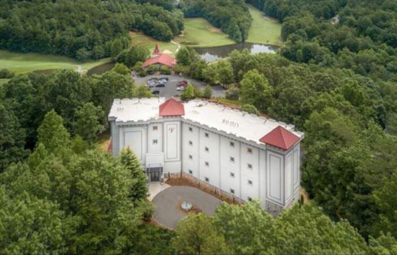 A large, white building with a red roof, surrounded by dense greenery and a small body of water.