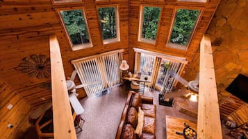 A cabin interior with a wooden ceiling, multiple windows, a central seating area with a brown leather sofa, and a stone wall.