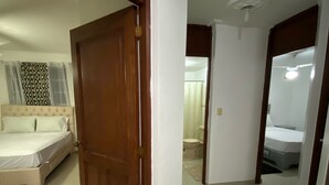 Quarto de hotel com uma cama grande, banheiro com chuveiro e corredor que dá para outro quarto.