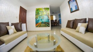 Uma sala de estar com dois sofás, uma mesa de centro de vidro, um quadro com uma paisagem de rio e um ventilador de teto.