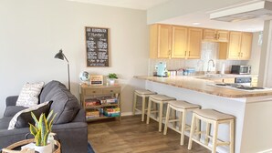 A modern kitchen with wooden cabinets, a central island with stools, and a dining area with a grey sofa and a globe decoration. - Cozy 2BR Lake Monroe Golf Condo IU Bloomington (Bloomington)