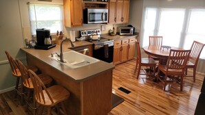 A kitchen with wooden cabinets, a central island with a sink, a stove, and a microwave. There are bar stools and a dining table with chairs. - Topsy's Trailer, Private, FAST WiFi, Small Dog Ok, HOT TUB, Close to AVL/Hendo (Arden)