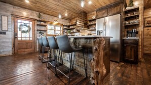 Une cuisine rustique aux murs et au sol en bois, avec un îlot central doté de tabourets de bar, un réfrigérateur en acier inoxydable et des étagères remplies d’ustensiles de cuisine.