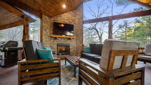 Une terrasse en bois avec un foyer en pierre, des sièges d’extérieur et un téléviseur fixé au mur.