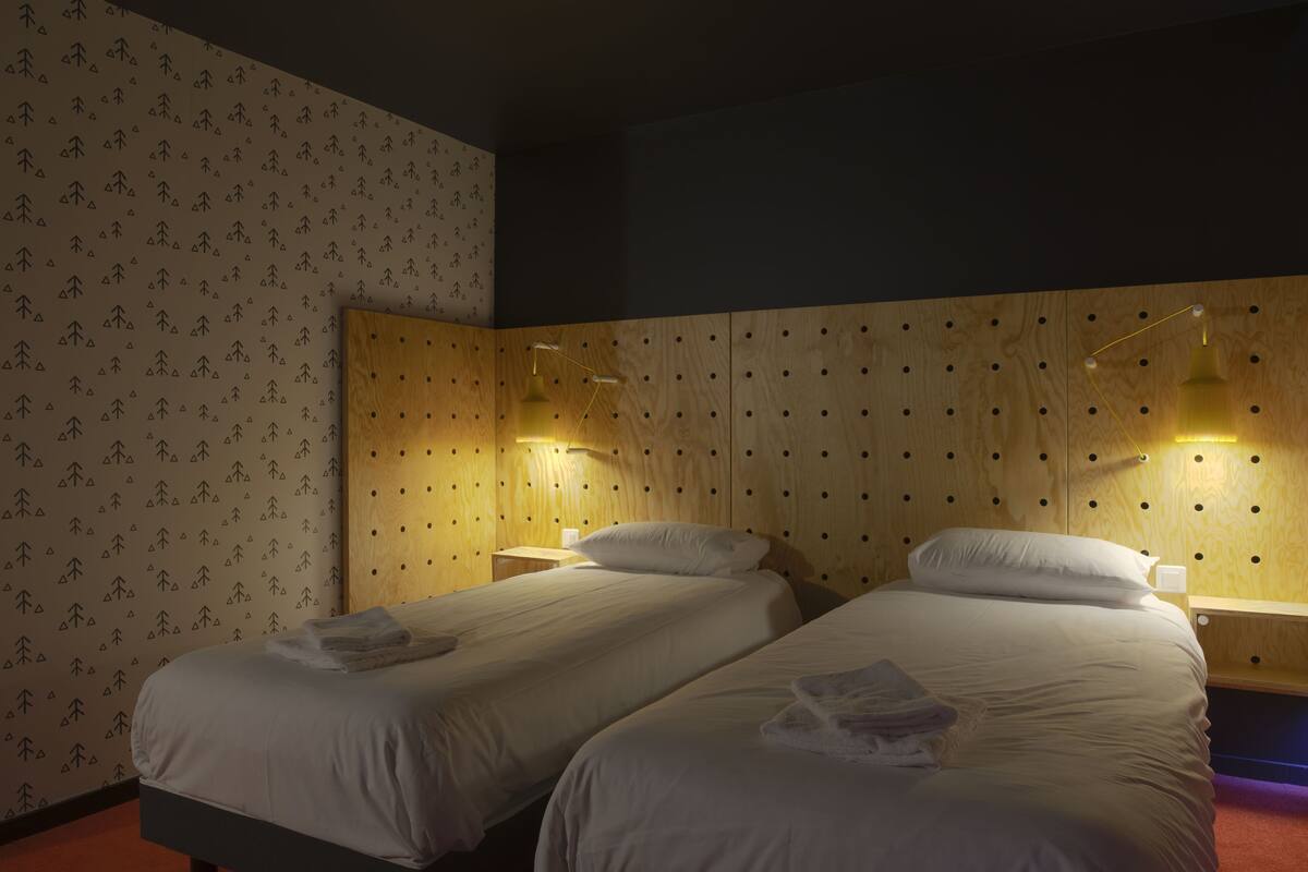 A hotel room with two single beds, wooden headboard with circular cutouts, and wall-mounted lamps.