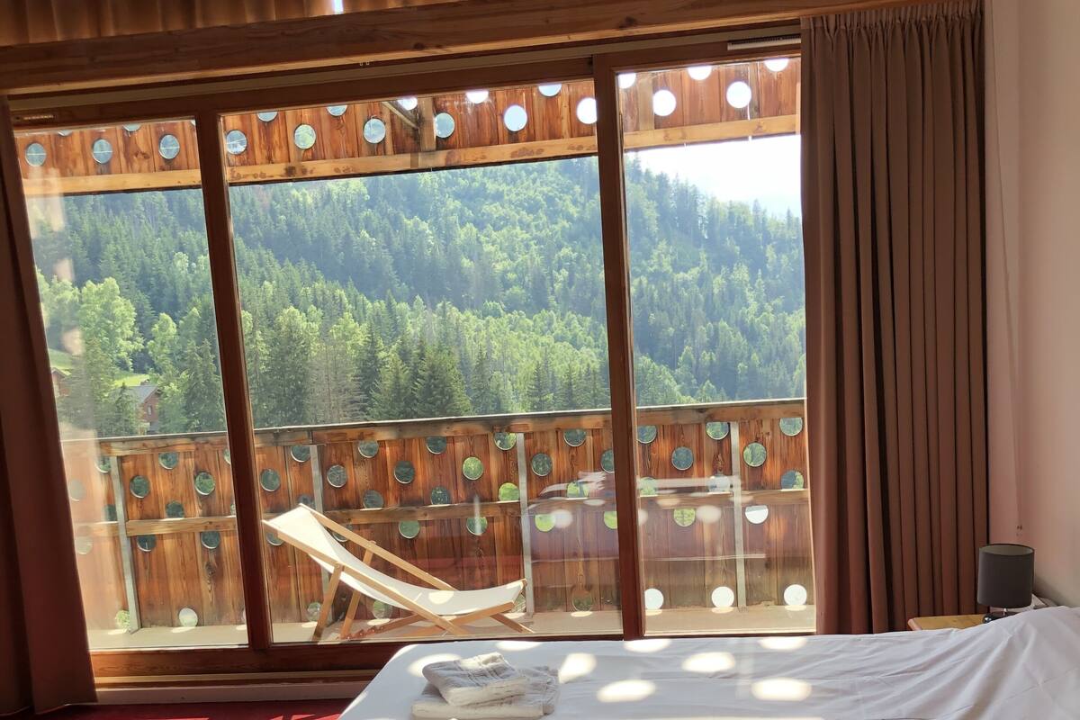 A bedroom with a large window, a bed with white linens, and a wooden deck with a chair.