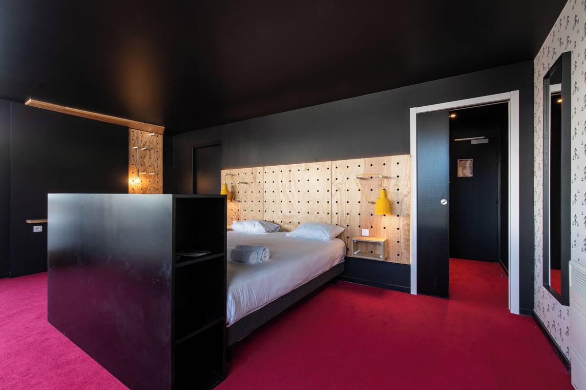 A modern bedroom with a bed, a wooden headboard, a red carpet, and a small bedside table with a lamp.