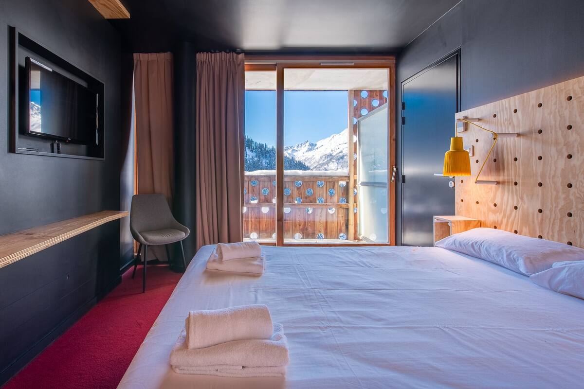 A hotel room with a large bed, a TV mounted on the wall, a wooden headboard, and a balcony with a mountain view.