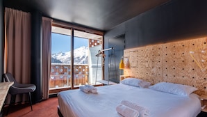A hotel room with a large bed, a desk, and a chair. There is a view of mountains through the window. - Timberlodge (Oz)