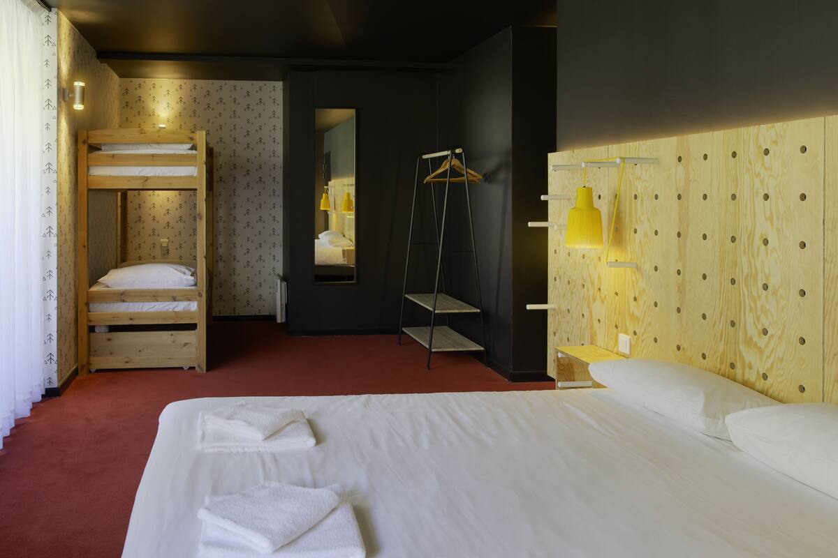 A hotel room with a bunk bed, a hanging clothes rack, a bedside table with a lamp, and a large bed with towels.