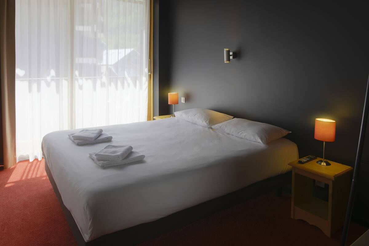 A neatly made bed with white linens and towels, two bedside lamps, and a view of the outdoors through sheer curtains.