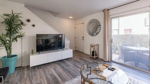 A modern living room with a flat-screen TV on a white console, a large potted plant, and a glass coffee table.