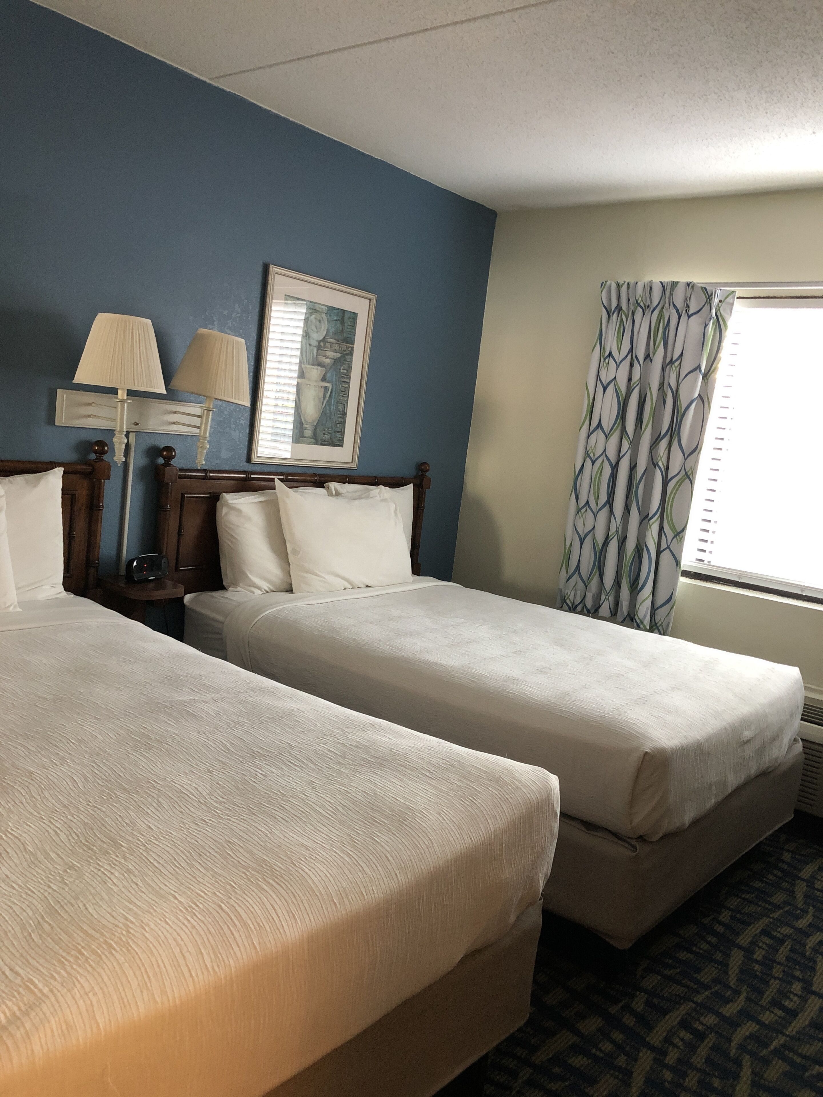 A hotel room with two single beds, a nightstand with a lamp, a framed picture on the wall, and a window with patterned curtains.
