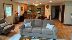 Ein geräumiges Wohnzimmer mit grauem Sofa, einem Holztisch und einem Essbereich, im Hintergrund ist eine Küche zu sehen.
