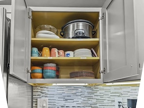 A kitchen cabinet with a crockpot, colorful plates, and storage containers.