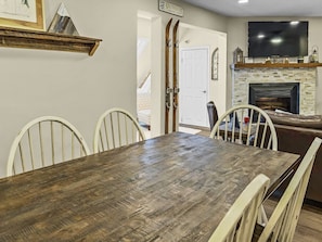 A dining area with a wooden table and white chairs, a fireplace, and a TV mounted above it.